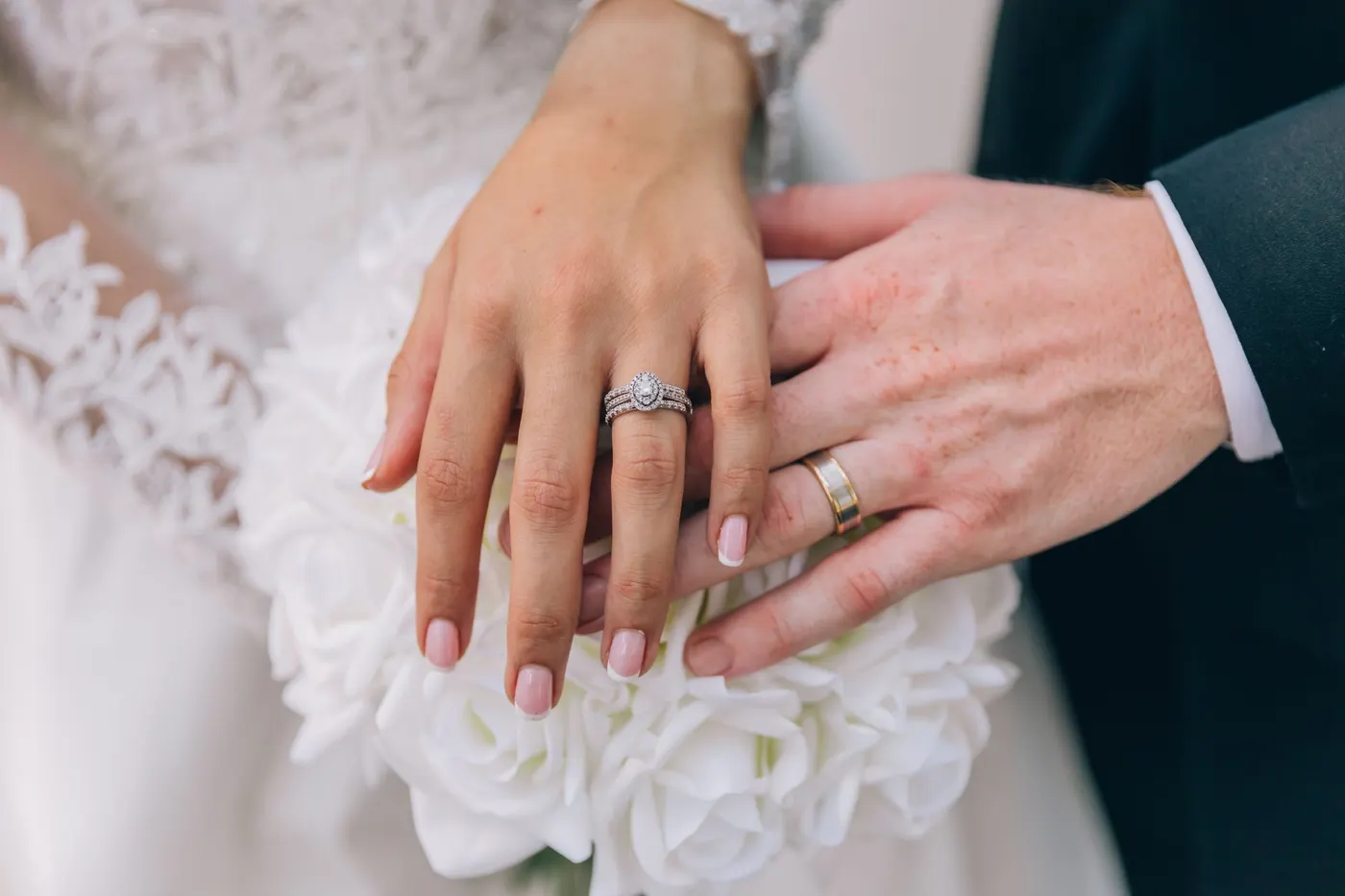 Mãos do casal com as alianças sobre um buquê de rosas brancas